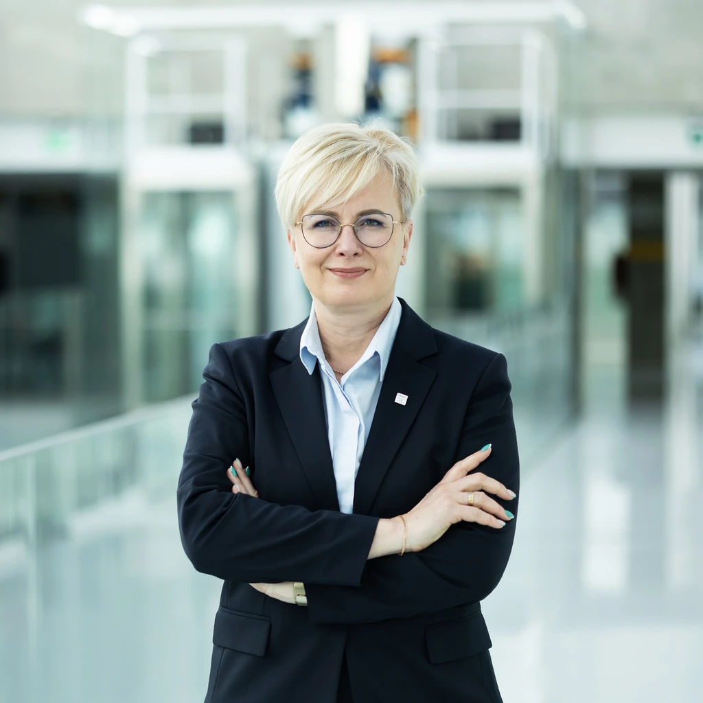 Portrait of Jurgita Stravinskienė, Head of the KTU Lifelong Learning Centre, photographed in the interior of Kaunas University of Technology&rsquo;s Santaka Valley &ndash; the science, studies, and business centre. A woman with short light hair and glasses looks directly into the camera with a subtle smile. She stands with her arms crossed, wearing a classic dark blazer and a light blue shirt.