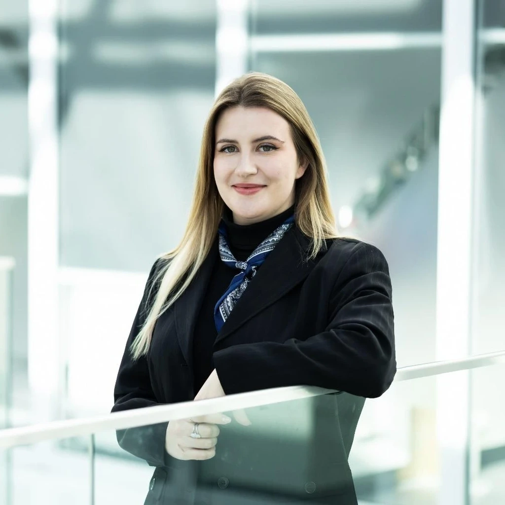 Portrait of Lukrecija A&scaron;embergaitė, Project Manager at the KTU Lifelong Learning Centre, photographed in a modern, bright university interior. A young woman with straight light hair and no glasses looks into the camera with a reserved smile. She is leaning on a modern glass railing, wearing a dark blazer and an ornamental scarf.