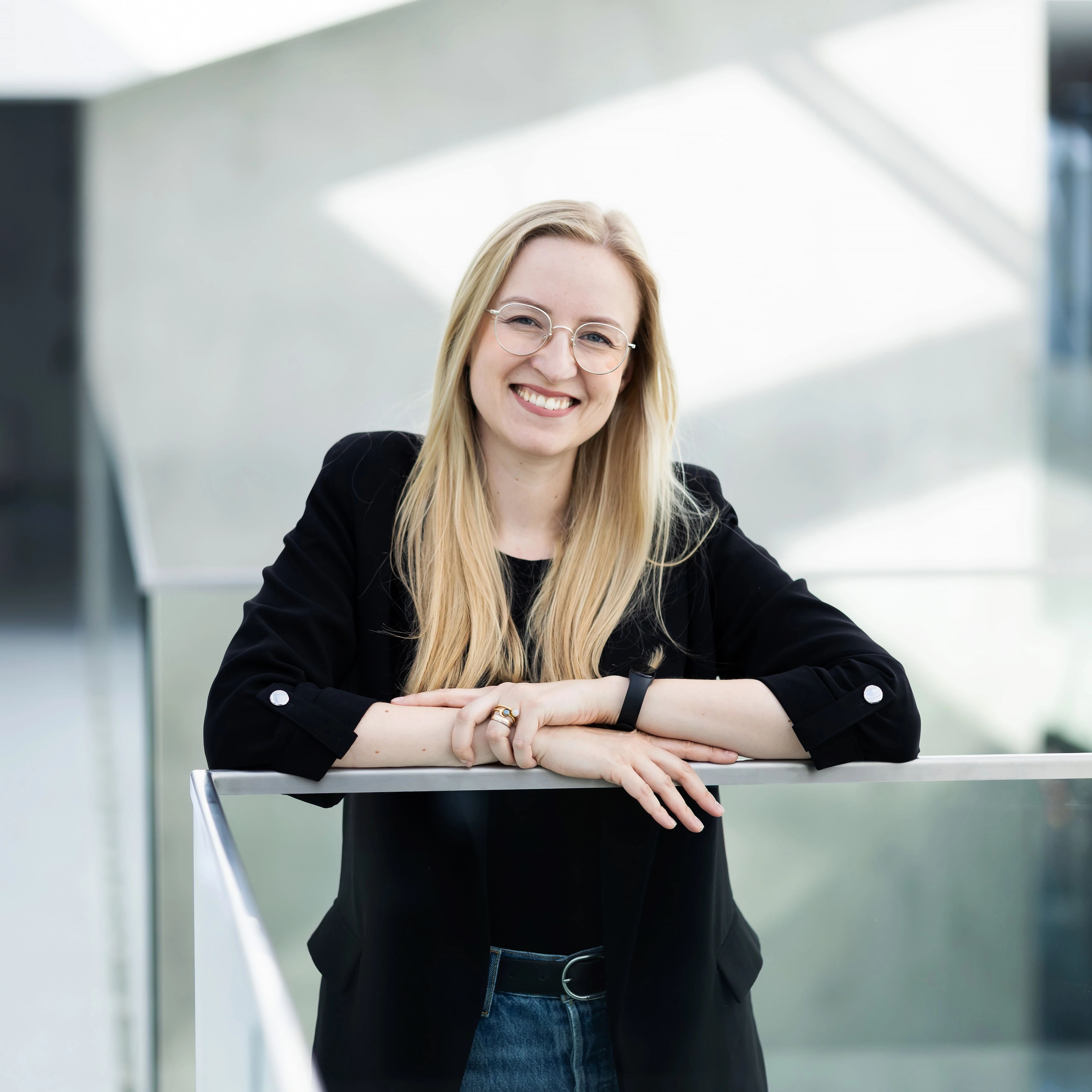 Portrait of Milda Guzaitytė-Raudonė, Business Client Project Manager at KTU, photographed in the interior of Kaunas University of Technology&rsquo;s Santaka Valley. A young woman with light, loosely falling hair and glasses looks directly into the camera and smiles. Leaning against a glass railing, she is wearing a black blazer and jeans.